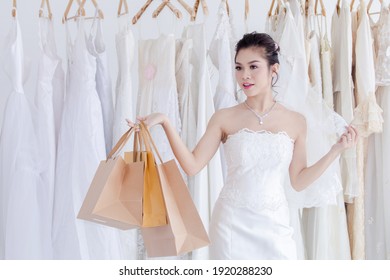 Beautiful Asian Happy Bride Holding Shopping Bags In Wedding Dress Studio, Young Modeling Present Shopping Bags In Front Of Many Wedding Dresses, Shopping For Engagement Or Wedding Ceremony Concept