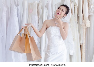 Beautiful Asian Happy Bride Holding Shopping Bags In Wedding Dress Studio, Young Modeling Present Shopping Bags In Front Of Many Wedding Dresses, Shopping For Engagement Or Wedding Ceremony Concept
