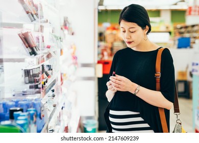 Beautiful Asian Korean Pregnant Woman Buying Cosmetic Product In Supermarket. Charming Young Future Motherhood Lady With Big Belly Holding Makeup Lipsticks And Choosing Color In Grocery Store.