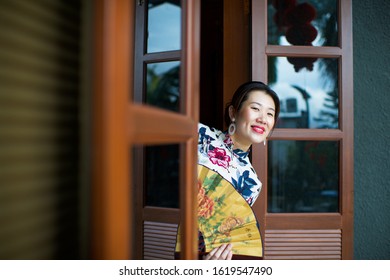 Beautiful Asian lady holding paper folding fan.Chinese female looking out window.Red lanterns reflection on window glass.Face with dimple.China town restaurant.Portrait of Oriental sexy model.