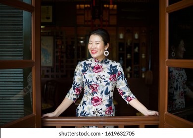 Beautiful Asian lady standing by window.Female wearing qipao.Cheongsam with flowers.Traditional eastern culture.Sexy model in restaurant.Oriental beauty.Face with dimples.Chinese New year greeting.