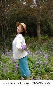 A Beautiful Asian Lady Standing In Spring Field Holding A Purple Bouquet Behind Her Back Smiling At Camera. 