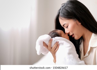 Weiße Hemdsfrau asiatische Mutter küsst ihr Neugeborenes im Schlafzimmer vor Glasfenstern mit weißem Vorhang, um Liebe und familiäre Bindungen zu zeigen.