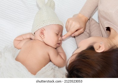 Beautiful Asian Mother Lying On Bed With Newborn Baby And Using Index Finger Touching On Baby Nose Newborn Gently While Toddler Sleeping On Bed. Adorable Infant Wearing Beige Knitted Hat.