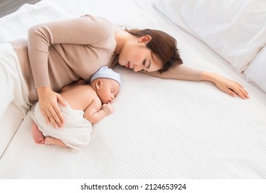 Beautiful Asian Mother Lying On Bed With Newborn Baby Wearing Blue Hat. In Home, Mom Hugging Little Baby With Love And Care. Woman Carying Her Adorable Infant While Toddler Deepy Sleeping In Bedroom.