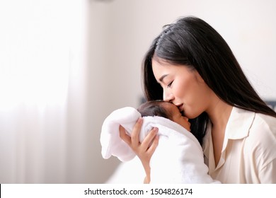 Beautiful Asian mother or mom is kissing at forehead of her newborn baby child in bedroom in front of glass windows with white curtain with concept love attractive and family bonding.