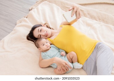 Beautiful Asian Mother Taking Selfie Picture By Smartphone With Daughter In Bed, Infant Lie Down On Mom Arm. Women And Baby Lie Down Together And Pay Attention With Camera. 