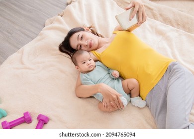 Beautiful Asian Mother Taking Selfie Picture By Smartphone With Daughter In Bed, Infant Lie Down On Mom Arm. Women And Baby Lie Down Together And Pay Attention With Camera. 