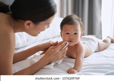 Beautiful Asian mother is wiping her newborn baby mouth gently by cloth diaper while the child is crawling on the bed. Touch of mother's love concept.