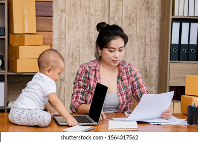 Beautiful Asian Mother Woman Taking Care Baby Multi Task Working At Home Using Laptop, Smartphone Packing Products Cardboard, Small Business Startup Entrepreneur, Freelance Lifestyle Shopping Online