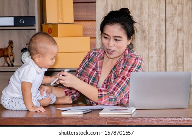 Beautiful Asian Mother Woman Taking Care Baby Multi Task Working At Home Using Laptop, Smartphone Packing Products Cardboard, Small Business Startup Entrepreneur, Freelance Lifestyle Shopping Online