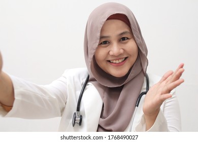 Beautiful Asian Muslim Female Doctor Wearing Hijab And Suit Taking Selfie Photo On Relax Time. Medical Doctor Doing Video Call, Smiling And Waving Saying Hi To Her Family From Her Workplace