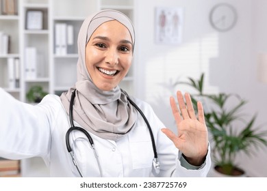 Beautiful asian muslim female doctor in hijab and suit taking selfie photo while resting. A doctor makes a video call, smiling and waving as he greets his family from his workplace - Powered by Shutterstock