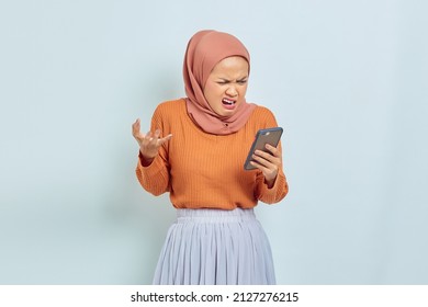 Beautiful Asian Muslim Woman In Brown Sweater And Hijab Holding Mobile Phone With Angry Expression Isolated On White Background. Muslim Lifestyle Concept