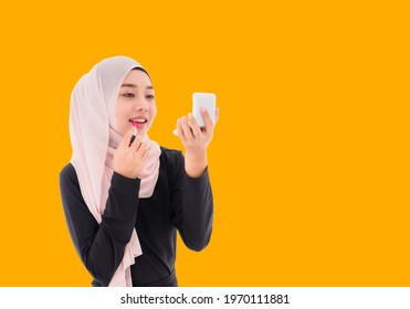 Beautiful Asian muslim woman in hijab standing holding mirror and applying pink lipstick on her lips isolated on yellow background. Happy young arabic female wearing headscarf and makeup by lipstick.