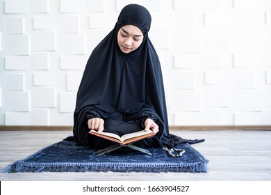 beautiful asian muslim woman reading the quran resting on rahel stand and turning page on the book, sitting on vintage prayer mat on knees, with misbaha resting on matt, bright cool tone background - Powered by Shutterstock