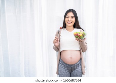 Beautiful Asian Pregnant Woman Eating Tomato With Fork Holding Bowl Of Salad Of Vegetables And Fruits, Healthy Having Lunch Standing Front Of White Curtain Background, Relaxing And Resting Living Room