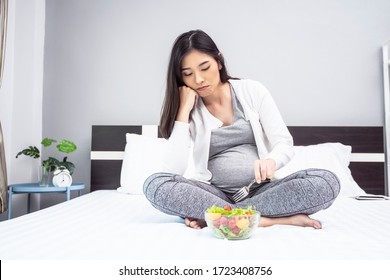 beautiful asian pregnant woman eating bowl of salad feeling sad on eating healthy and stress out from pregnancy, sitting on bed with glass bowl of vegetables and fruit using a fork to pick up the food