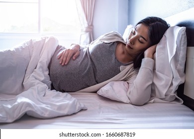 Beautiful asian pregnant woman laying down on bed sideways sleeping taking a nap with pregnant belly out, resting and relaxing in bed room from hormone stress, comfy wearing stretch pants and cardigan - Powered by Shutterstock