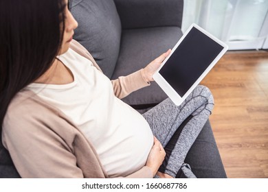 wunderschöne asiatische schwangere Frau, die mit einem intelligenten Tablet-Gerät lächelnd sieht Video, die Hände auf den Babyklumpen, sitzend auf Sofa entspannend Ruhen im Wohnzimmer mit Backsteinstruktur Wand und weißen Vorhängen