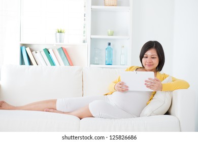 Beautiful Asian  pregnant woman using tablet computer sitting on sofa at home - Powered by Shutterstock