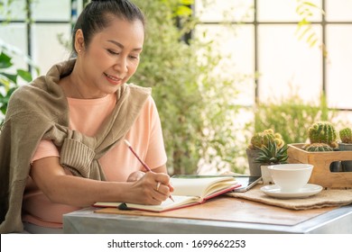 Beautiful Asian senior woman writing diary or planning in notebook at home greenhouse garden. Smiling retired woman relaxing at home during quarantine. Elderly lifestyle and healthcare concept.