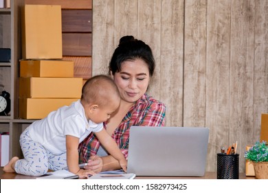 Beautiful Asian Single Mother Taking Care Baby Multi Task Working At Home Using Laptop, Smartphone Packing Products Cardboard, Small Business Startup Entrepreneur, Freelance Lifestyle Shopping Online