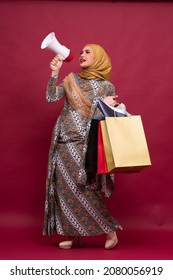 Beautiful Asian Woman In Batik Dress And Hijab Holds In Hand Bullhorn Public Address Loud Hailer With Shopping Bags On Red Background. Scream In Megaphone. Business Shopping Sales Lifestyle Concept.