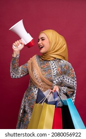 Beautiful Asian Woman In Batik Dress And Hijab Holds In Hand Bullhorn Public Address Loud Hailer With Shopping Bags On Red Background. Scream In Megaphone. Business Shopping Sales Lifestyle Concept.