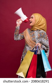 Beautiful Asian Woman In Batik Dress And Hijab Holds In Hand Bullhorn Public Address Loud Hailer With Shopping Bags On Red Background. Scream In Megaphone. Business Shopping Sales Lifestyle Concept.