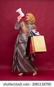 Beautiful Asian Woman In Batik Dress And Hijab Holds In Hand Bullhorn Public Address Loud Hailer With Shopping Bags On Red Background. Scream In Megaphone. Business Shopping Sales Lifestyle Concept.