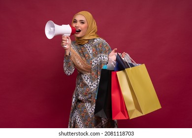 Beautiful Asian Woman In Batik Dress And Hijab Holds In Hand Bullhorn Public Address Loud Hailer With Shopping Bags On Red Background. Scream In Megaphone. Business Shopping Sales Lifestyle Concept.