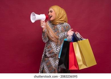 Beautiful Asian Woman In Batik Dress And Hijab Holds In Hand Bullhorn Public Address Loud Hailer With Shopping Bags On Red Background. Scream In Megaphone. Business Shopping Sales Lifestyle Concept.