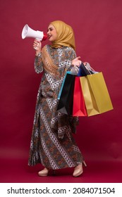 Beautiful Asian Woman In Batik Dress And Hijab Holds In Hand Bullhorn Public Address Loud Hailer With Shopping Bags On Red Background. Scream In Megaphone. Business Shopping Sales Lifestyle Concept.