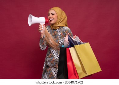 Beautiful Asian Woman In Batik Dress And Hijab Holds In Hand Bullhorn Public Address Loud Hailer With Shopping Bags On Red Background. Scream In Megaphone. Business Shopping Sales Lifestyle Concept.