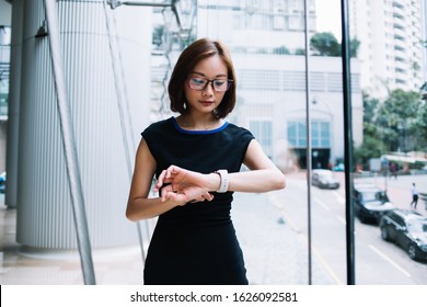 Beautiful Asian Woman In Black Dress And Glasses Checking Time On Smart Watch While Standing Near Window In Modern Office Building