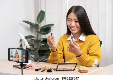 Beautiful asian woman blogger using camera phone recording vlog video live streaming and applying makeup for review cosmetics at home.