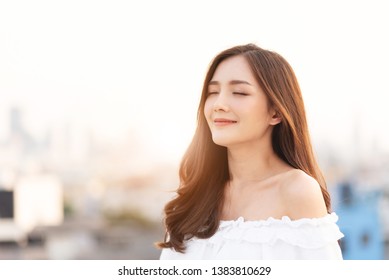 Beautiful Asian Woman Is Breathing Fresh Air. Smiling Female Is Standing On Top Of House Roof Over City Background At Outdoor. Relaxation, Eyes Closed.