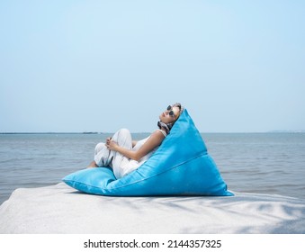 Beautiful Asian Woman In Casual White Shirt And Trousers Wearing Sunglasses And Hair Scarf Reclining On Blue Bean Bag Seat On The Big Rock With Sea View And Blue Sky Background On Sunny Day In Summer.