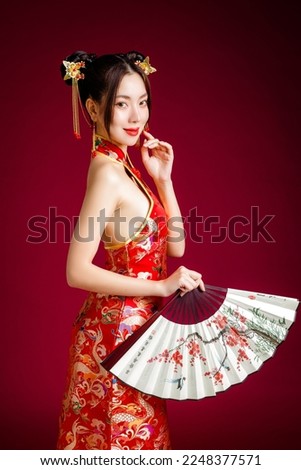Beautiful Asian woman with clean fresh skin wearing traditional Chinese dress holding fan posing on red background. Chinese text means looking for spring and happy to see plum.