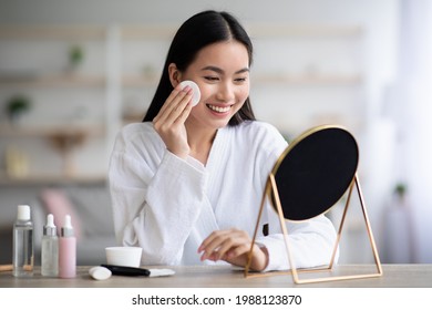 Beautiful asian woman cleaning her face, using cotton pads and cleansing product, looking at mirror in bedroom. Young attractive korean lady using face toner and cotton pad, home interior, empty space