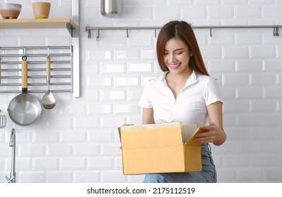 Beautiful Asian Woman Customer Receive Post Shipment Parcel At Home. Japanese Girl Opening Cardboard Box While Seated On Countertop In White Modern Kitchen. Online Shopping Order, Delivery Concept