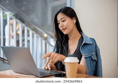 A beautiful Asian woman in a denim jacket is focused on her work, wearing earphones while working remotely on her laptop from a coffee shop. businesswoman, freelance, college student - Powered by Shutterstock