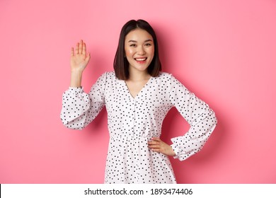 Beautiful Asian Woman In Dress Saying Hello, Waving Hand To Greet And Say Hi, Smiling Friendly At Camera, Standing Over Pink Background