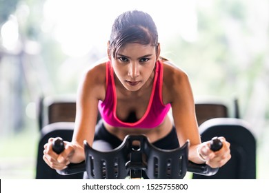 Beautiful Asian Woman Exercising On Stationary Cycling Machine In Indoor Fitness Gym, Determination Face. Sport Recreational Activity, People Workout, Or Healthy Lifestyle Concept