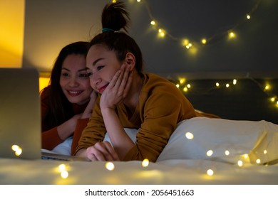 Beautiful Asian Woman Friends Lying On The Bed In Bedroom Watching Movie Or Online Shopping Together On Laptop Computer. Modern Female Enjoy Weekend Activity Lifestyle With Technology Together At Home