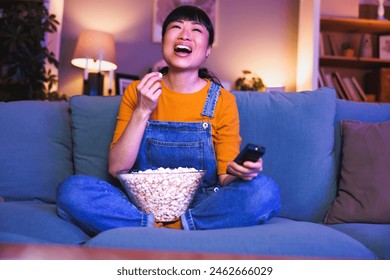 Beautiful Asian woman having fun sitting on living room couch switching channals using remote control and relaxing at home late at night watching TV - Powered by Shutterstock