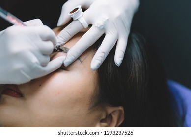 Beautiful Asian Woman Having Professional Eyebrow Tattoo In Beauty Salon. Cosmetologist Applying Permanent Make Up On Eyebrows.