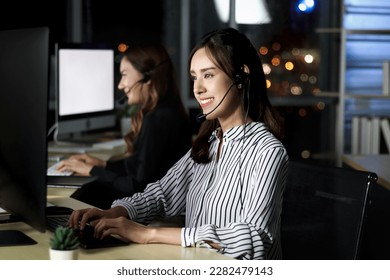 Beautiful Asian woman with headphones work night shift at call center customer care service desk, looking at desktop computer, typing answer question, female with hands-free phone work hard overtime. - Powered by Shutterstock