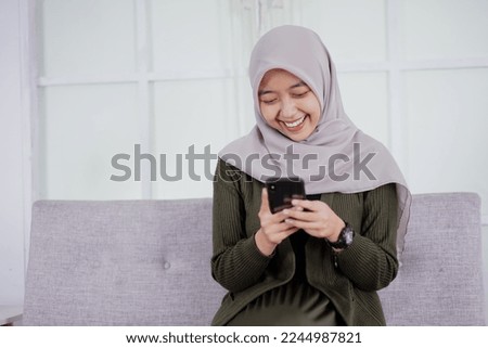A beautiful Asian woman in hijab smiles cheerfully sitting on a sofa using a mobile phone, receiving good news